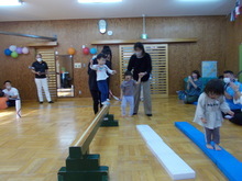 室内の広いスペースで、複数の子どもが保護者に支えられながら平均台を渡る種目をしている様子が写された写真
