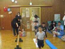 室内の広いスペースで、複数の子どもが保護者に支えられながら平均台にチャレンジしており、周囲には座って見守る人々がいる様子が写された写真