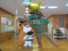 トナカイの着ぐるみを着た人物とクリスマスツリーの帽子をかぶった人物が並び、前には白と緑の服を着た子どもが立っている写真