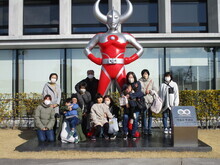 屋外に立つ巨大なキャラクターの像の前で、複数名の大人と子どもが集まり、記念撮影をしている様子の写真