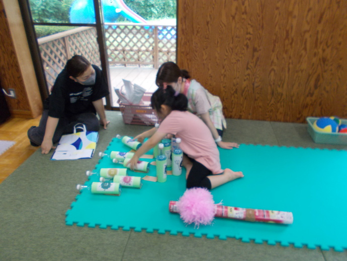 室内のスペースで、緑色のジョイントマットの上に複数のペットボトルを並べたボウリングを、複数の人がそれを手に取って遊んでいる様子の写真