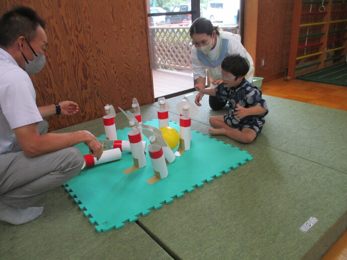 緑のジョイントマット上にペットボトルを使った赤白の手作りボウリングピンが並び、黄色いボールを使って遊んでいる写真