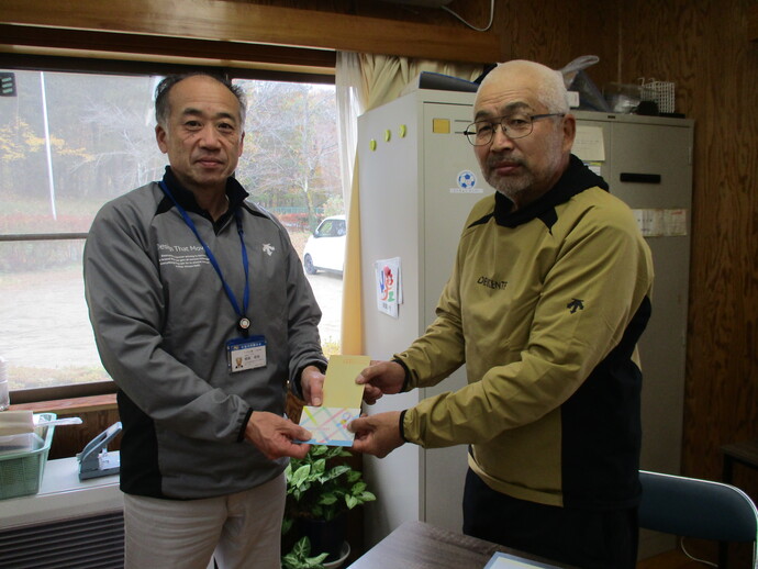 屋内の窓際で、グレーのジャンパーを着て名札を下げた男性と、眼鏡をかけ黄土色のウェアを着た白髪混じりの吉田様が両手で黄色い封筒と色鮮やかなカードに両手で持っている贈呈式の記念写真