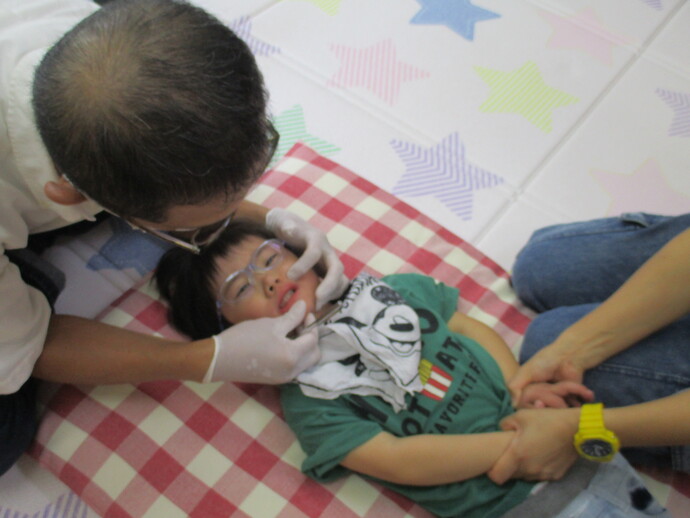 緑色のシャツを着た子どもが仰向けになり、隣で別の人物が子どもの腕を優しく支えて歯科検診をしている様子を写した写真