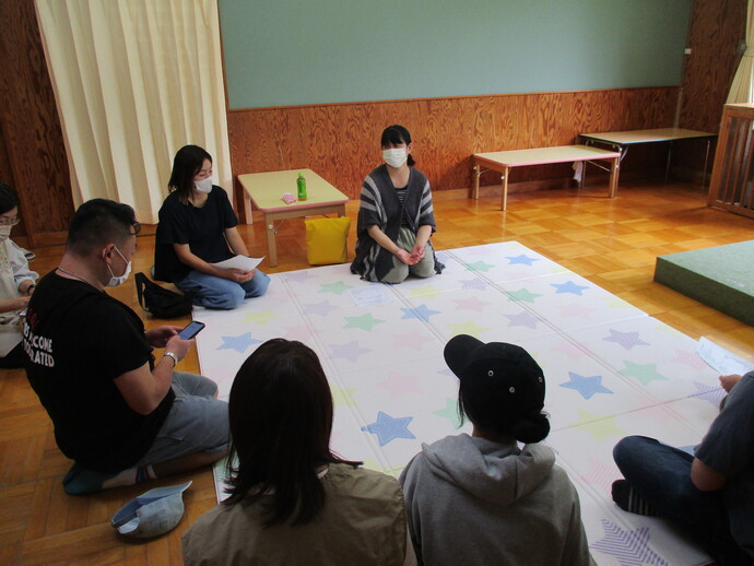 室内で床に敷かれた星柄の大きなシートの上に複数人が円形に座り、手元に紙を持ちながら話している保護者研修会の様子を写した写真