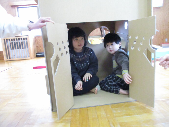 箱の前面には開いた扉と丸い穴があるダンボールハウスの中に二人の子どもが座っている写真