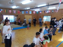 室内の広いスペースで、国旗が天井に並び、複数の大人と子どもが集まり、開会式をしている様子が写された写真