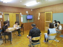 木の壁の室内で、中央に椅子が並び、複数の人物が座って医療的ケアに関する講義を受けており、前方では机にパソコンと人形が置かれ、壁にはモニターが設置されている写真