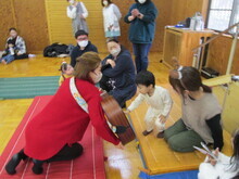 赤い服の人物がギターを持ち、白い服の子どもが近づいて触れようとして、周囲には数人が座って見守っている写真