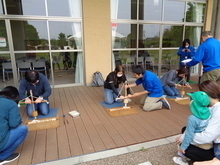 屋外のウッドデッキで複数の人が膝をつき、木の板の上に置かれた器具を使って火起こし体験をしている写真