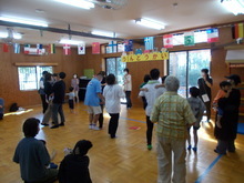 室内の広いスペースで、壁には「うんどうかい」と書かれた飾りがあり、複数の大人と子どもが集まって体操をしている様子が写された写真