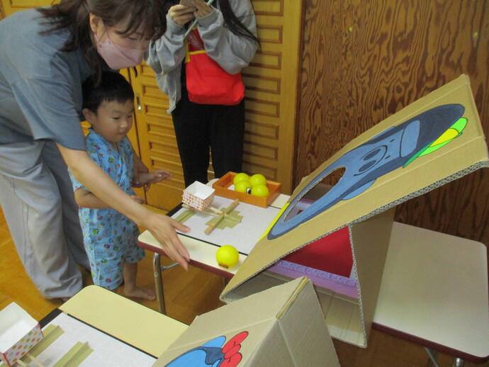 テーブル上に段ボールで作られた傾斜台があり、キャラクターの顔に穴が開いた的当てのような仕掛けが描かれ、子どもがボールを使った遊びに参加している様子の写真