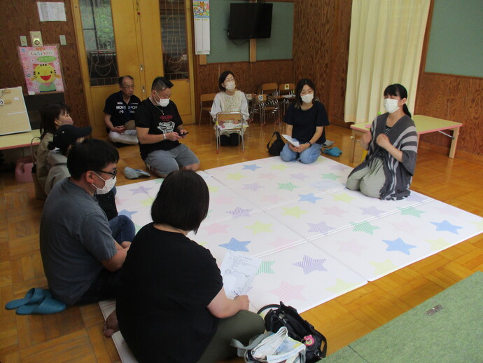 室内で床に敷かれた星柄の大きなシートの上に複数人が円形に座り、手元に紙を持ちながら右手に座っている女性の話を聞いている保護者研修会の様子を写した写真