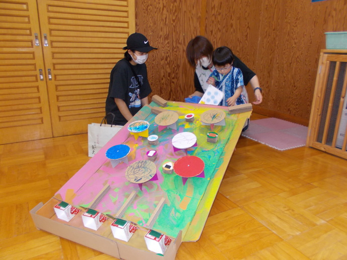 室内で、床に置かれた大きな段ボール製のボールころがしの工作物があり、斜面にピンクや緑の絵の具で彩色され、複数の円形や四角形のパーツが取り付けられた構造になっており、周囲には3人が座ってその作品を見たり操作したりしている様子がある写真