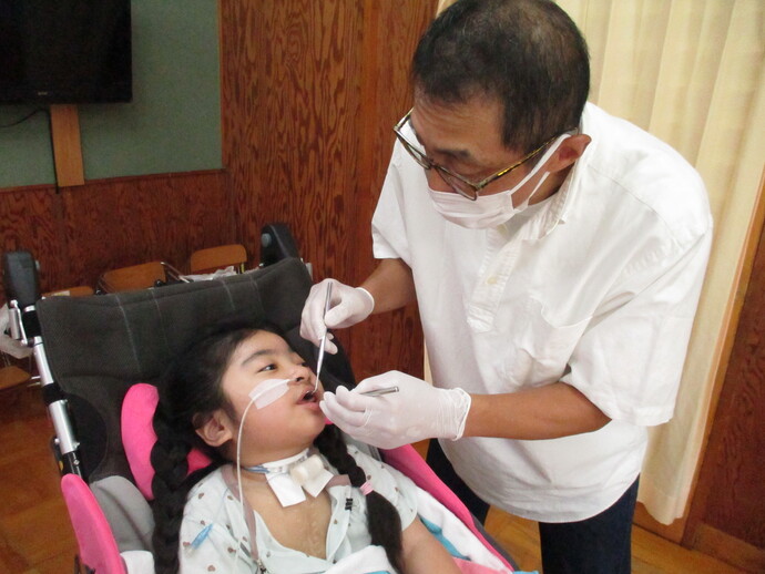 車椅子に座った子どもの胸元に医療用チューブがあり、白い手袋をした人物が歯科検診をしている様子を撮影した写真