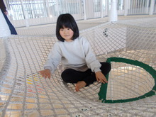 明るい屋内の遊び場に設置された白い網状の巨大な遊具の中に、女の子が笑顔で座っている様子の写真