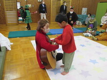 赤い服の人物がギターを抱え、赤い服と緑のズボンを履いた子どもが弦に触れて交流している写真