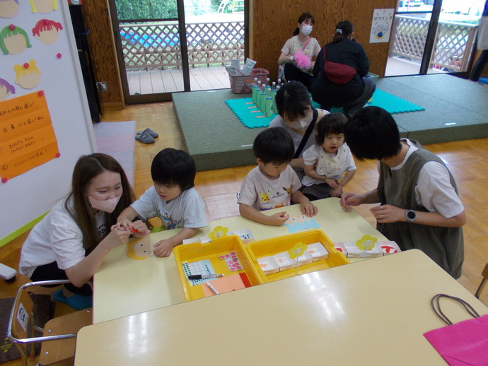 室内のスペースで、テーブルに黄色のトレーと紙工作の材料が並び、複数の子どもが座ってお面づくりの製作活動をしている写真