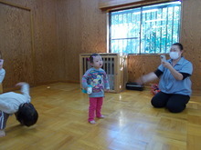 室内で中央にカラフルな服を着た幼児と左側に別の子どもが曲に合わせてダンスをし、右側ではマスクを着けた大人が床に座ってカメラを構えている様子が写された写真