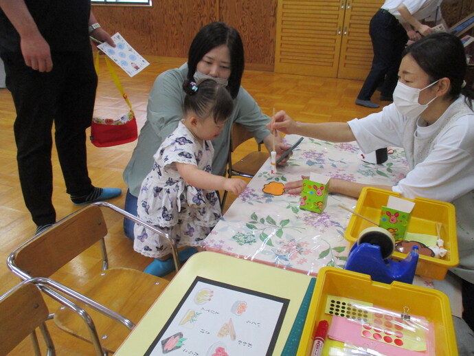 花柄のビニールクロスの上に色紙やのり、テープなどの工作道具が並び、子どもが大人と一緒に紙工作をしている様子の写真