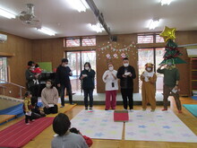 広間にクリスマスツリーやトナカイの仮装などをした関係者が横一列に並んで話をしているクリスマス会の様子を捉えた写真