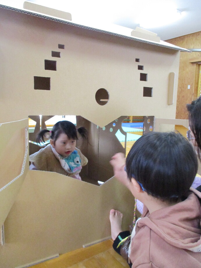 ダンボールハウスの中央の大きな開口部から子どもが中にいて外側の子どもが手を伸ばしている写真