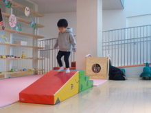 床に赤と緑の三角形のクッション遊具が置かれ、その上を灰色のパーカーと黒いズボンを着た子どもが歩いて降りている様子が写された写真