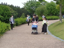 緑豊かな公園の舗装された遊歩道で、中央にベビーカーを押す人と隣を歩く人がおり、周囲には散歩を楽しむ複数の人々がいる写真