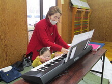 テーブル上に置かれた電子ピアノを赤い服の人物が演奏し、前には小さな子どもが鍵盤に手を伸ばしている様子が見える写真