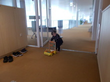 廊下ような場所で、幼い子どもが黄色い押し車のおもちゃに手をかけ歩いている様子の写真