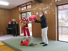 赤いサンタクロース衣装を着た人物がギターを持ち、別の人物が赤い箱を掲げ、床には赤い服の子どもが横たわりっている様子が写された写真