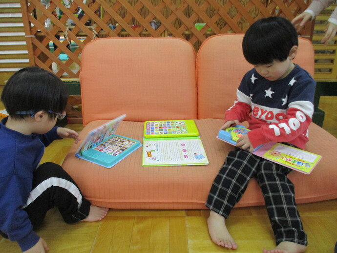 二人の子どもが床に座ったりソファに腰掛けたりしながらカラフルな絵本や知育本を広げて読んでいる室内の写真