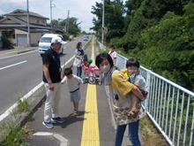黄色の誘導ブロックが敷かれた歩道で、一人の女性が幼児をおんぶし、他の大人が子どもと手を繋いでいる様子の写真
