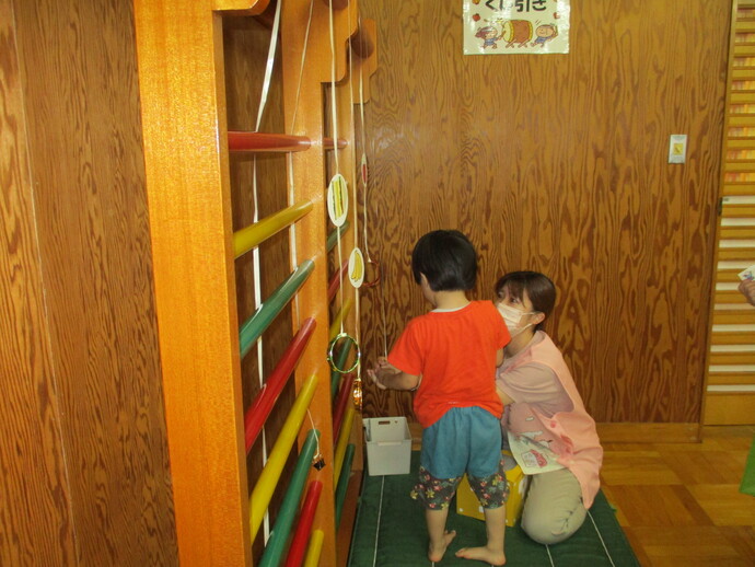 木製の壁際に設置された赤・黄・緑の横棒が並ぶ遊具に、複数の輪が紐で吊るされており、子どもがその輪に触れながら遊んでいる様子の写真