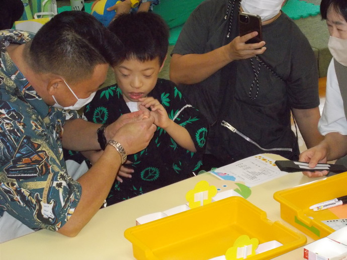 テーブルの上に黄色のトレーと紙工作の材料が並び、大人が子どもの手を取りながらお面づくりをしている室内の写真