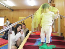 赤いカーペットの階段で白い服を着た子どもが黄色と緑の布を体にまとい、手すりのそばに座る人物がスマートフォンを構えている写真