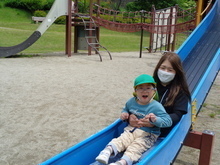 青い滑り台の上で、緑色の帽子をかぶった子どもが前に座り、その後ろに大人が一緒に滑っている様子が写っている屋外公園の写真