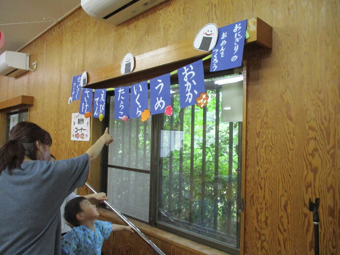 窓の上に「おにぎりのおめんをつくろう」と書かれた青い旗が並び、梅や鮭などの具材イラストが描かれた装飾が吊るされ、来場者の親子連れが具材を選んでいる様子の写真