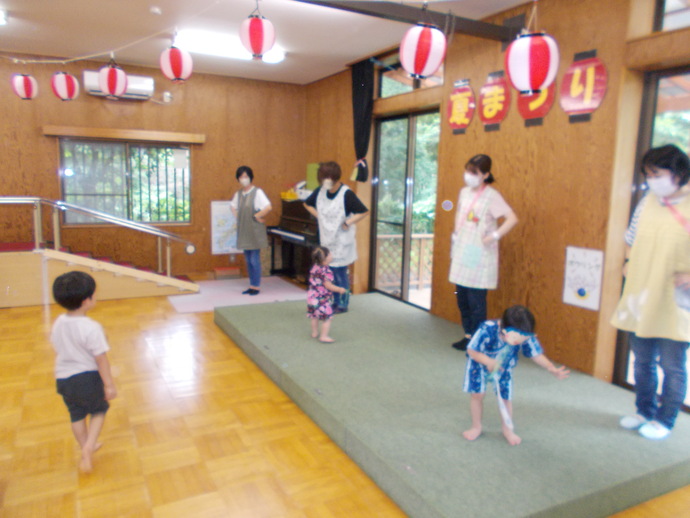 室内のスペースで奥の壁には「夏まつり」と書かれた飾りがあり、子供たちと複数のエプロンを着た人物が「はたらく細胞」体操をしている写真