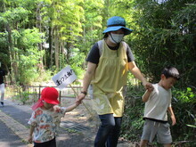 屋外の舗装された小道で、水色の帽子とエプロン姿の職員が、2人の幼い子どもを両手で引いて歩いている写真