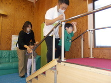 室内で小さな階段と手すりの上で前方に子どもが手すりを握って登り、後方では別の子どもが保護者に支えられている様子が写された写真