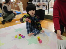 室内で星柄のシートの上に座る子どもが、カラフルなボールを並べて遊んでおり、周囲には赤い服の人物の腕や他の人々が見える写真