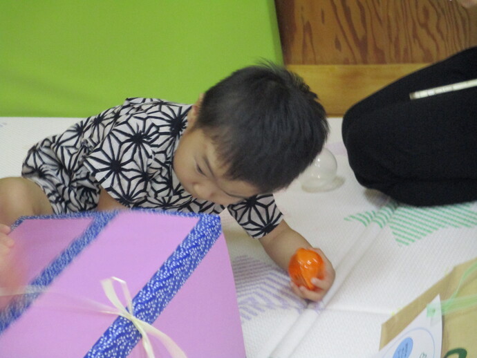 白地に黒い幾何学模様の浴衣を着た子どもが床に座り、紫色の箱に青い模様のリボンがかけられた箱から右手でオレンジ色の小さな玉を出しくじ引きを楽しんでいる写真