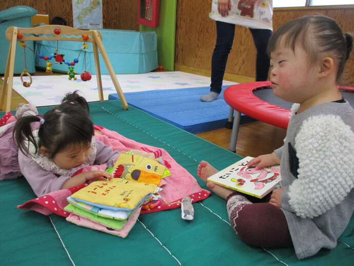 マットの上で一人の幼い子どもが布絵本を重ねてうつ伏せで見ており、もう一人の子どもが座って厚紙絵本を手に持っている写真