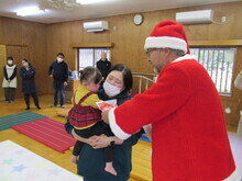 サンタクロースに扮した男性が、母親らしき女性に抱っこされ女の子に、プレゼントを手渡しているクリスマス会の様子の写真