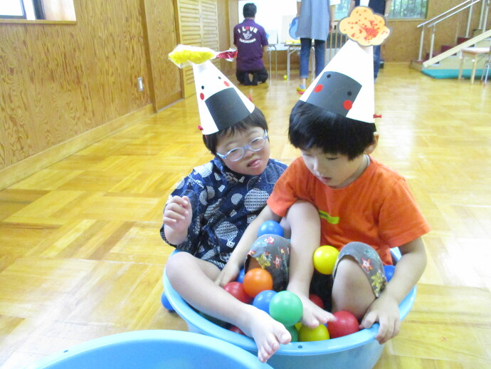 子どもがカラフルなボールが入った青いプラスチック容器に座り、頭にはおにぎりを模した紙製の帽子をかぶり、帽子には梅や鮭などの具材イラストが飾られている様子の写真