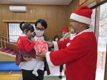 赤いサンタクロース衣装を着た人物がプレゼントを手渡し、別の人物が子どもを抱いて受け取っている様子が写された写真