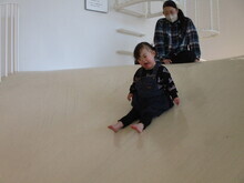 白く緩やかな傾斜のある大きな室内遊具の斜面を、幼い女の子が裸足で滑り降りており、その後ろの斜面の上で女性が見守っている様子の写真