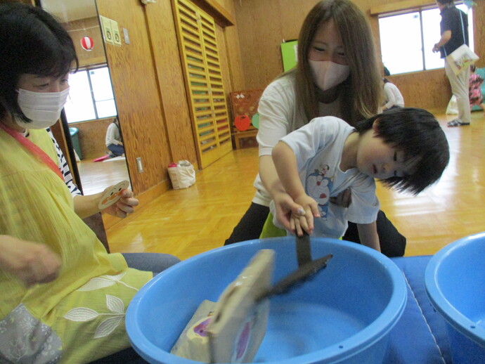 室内の明るい部屋で、青い大型のたらいに浮かぶ木製の船を子どもが道具でおもちゃすくいを楽しんでいる写真