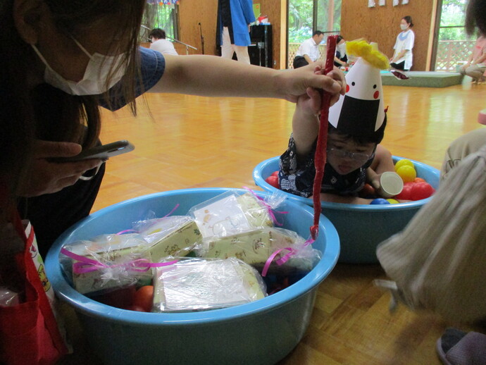 プラスチック容器が二つ置かれ、一方にはカラフルなボールと紙製のおにぎり帽子をかぶった子どもが入り、もう一方にはリボンで結ばれた複数の包装品が詰められており、近くで大人が赤い棒を持って遊びを補助している様子の写真